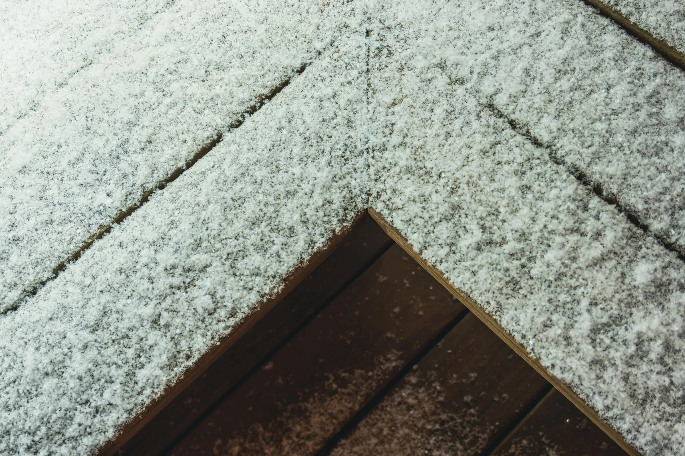 Close-up of snow-covered concrete pavement with a dark wooden object or trim visible at the bottom edge.